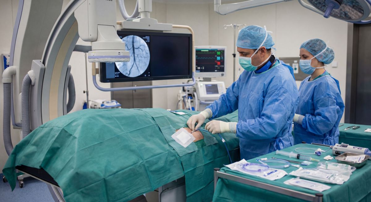 Badanie angiograficzne w trakcie – personel w sterylnych strojach pracuje przy pacjencie na stole, obok ramię C aparatu RTG i monitory z obrazem naczyń oraz parametrami życiowymi.