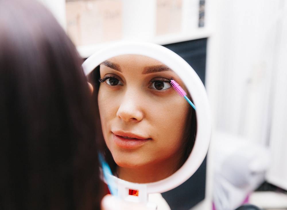 kobieta nakładająca odżywkę na rzęsy i patrząca w lustro