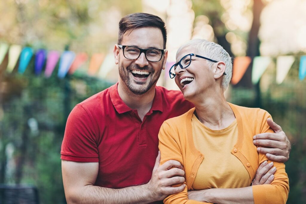 Uśmiechnięty mężczyzna w czerwonej koszulce polo i okularach obejmuje roześmianą kobietę z krótkimi siwymi włosami w okularach; w tle kolorowe girlandy na świeżym powietrzu