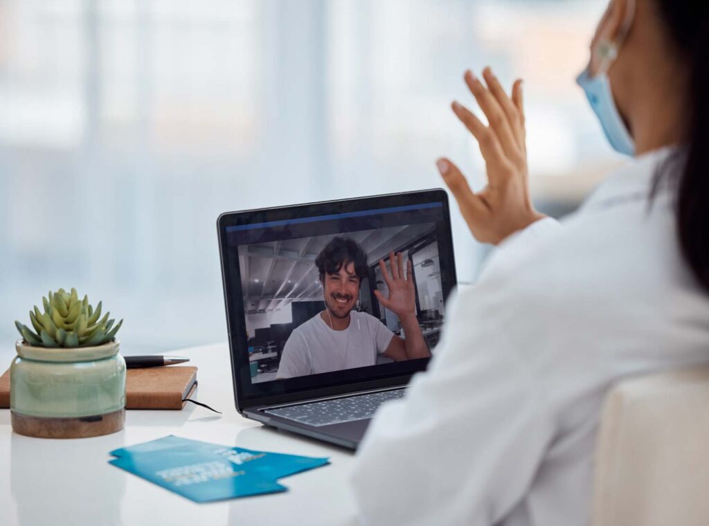 Widok zza ramienia lekarki w maseczce, prowadzącej e-wizytę na laptopie. Na ekranie widać uśmiechniętego pacjenta ze słuchawkami, z którym lekarka wymienia powitalne machnięcie ręką.